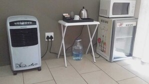 Una habitación con un ventilador de pie, una mesita con una tetera, una botella de agua y un refrigerador.