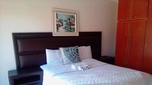 Una cama bien tendida con sábanas blancas y una almohada decorativa, un cuadro en la pared y un armario a la derecha.