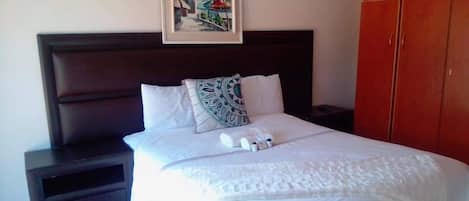 A neatly made bed with white linens and a decorative pillow, a framed picture on the wall, and a wardrobe to the right.