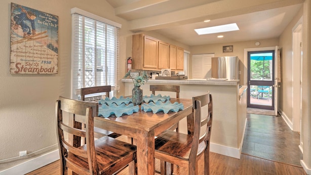 A dining area with a wooden table and chairs, a kitchen in the background, and a door leading outside.