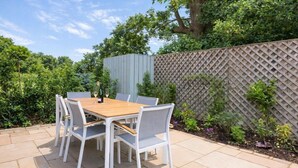 Een terras met een houten tafel en stoelen, omringd door een haag en een lattenhek.