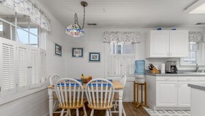 A kitchen with white cabinets, a wooden dining table with two chairs, a hanging light fixture, and a window with shutters. - Perfect beach cottage for your Lewes vacation! 10 Texas (Lewes)
