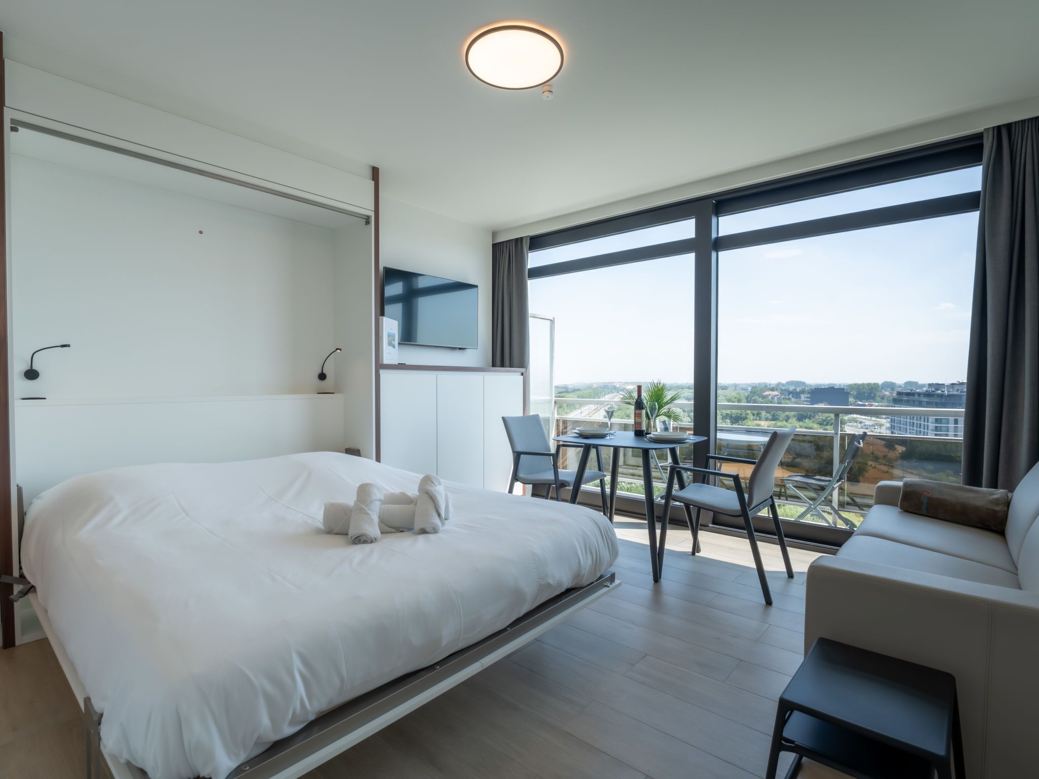 Ein modernes Hotelzimmer mit Bett, Fernseher, Balkon mit Tisch und Stühlen sowie Blick auf die Stadt.