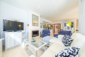 Een woonkamer met een televisie, witte meubels, een glazen salontafel en een open haard.