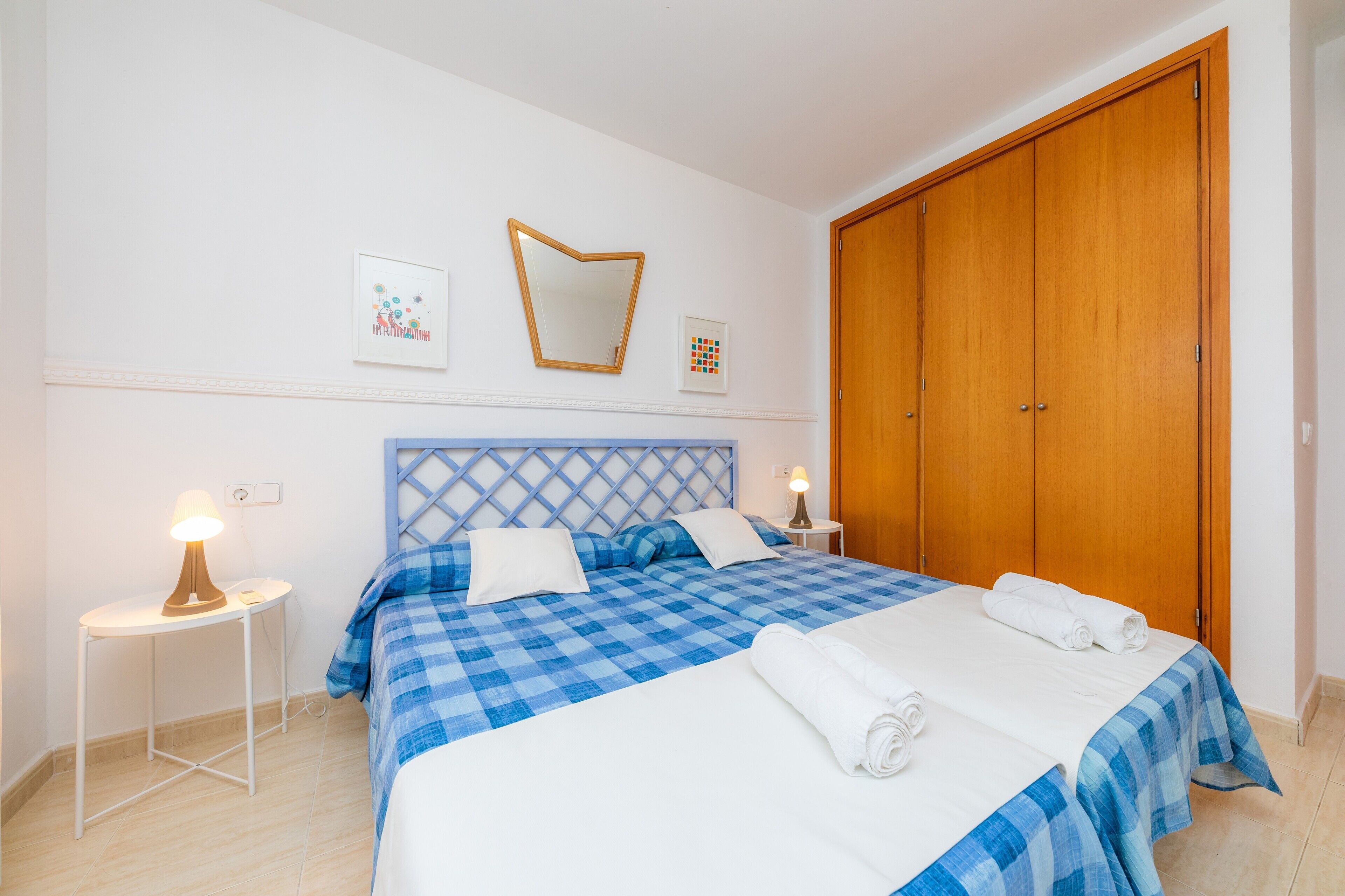 A neatly made bed with a checkered blue and white bedspread, two white pillows, and rolled towels on the bed. There is a wooden wardrobe to the right and a small bedside table with a lamp on the left.