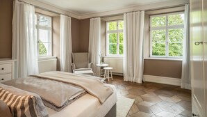 A bedroom with a bed, two chairs, a small table, and large windows with curtains. - Schloss Kirchberg – Apartment Rosé (Immenstaad)