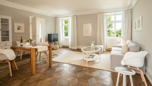 A modern living room with a wooden herringbone floor, a dining table with chairs, a sofa, a TV, and a window with curtains. - Schloss Kirchberg – Apartment Rosé (Immenstaad)