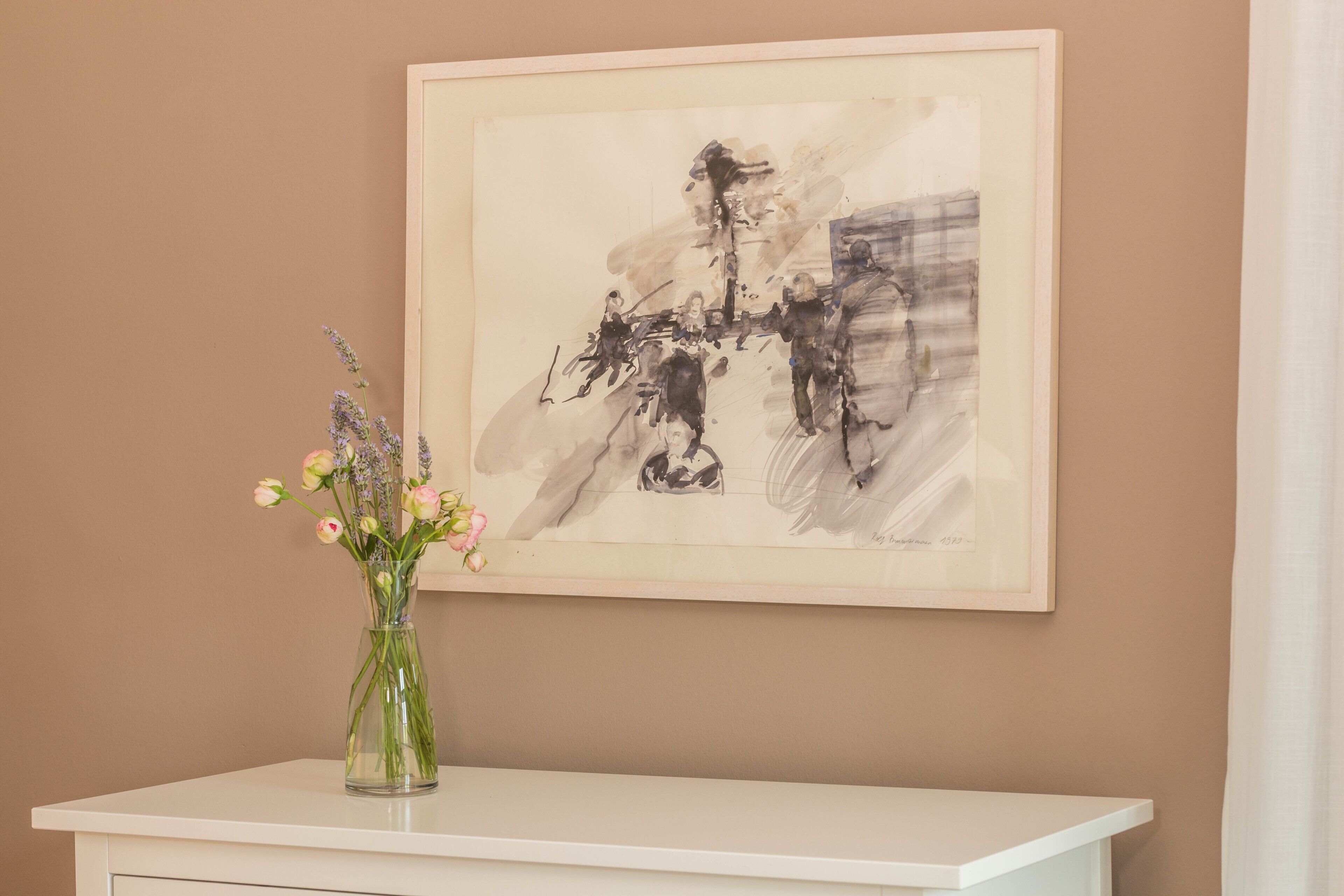 Ein gerahmtes, abstraktes Gemälde an einer Wand, eine Vase mit Blumen auf einer weißen Oberfläche darunter und eine schlichte Wand.