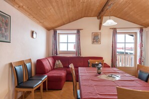 A cozy room with a wooden ceiling, a red sofa, a dining table with chairs, and two windows with curtains.