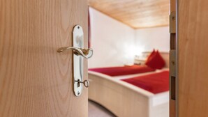 A wooden door with a metal handle and a keyhole, leading to a room with a bed and a red blanket.
