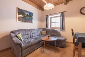 A living room with a grey sofa, a round wooden coffee table, a painting on the wall, a window with curtains, and a wooden ceiling.