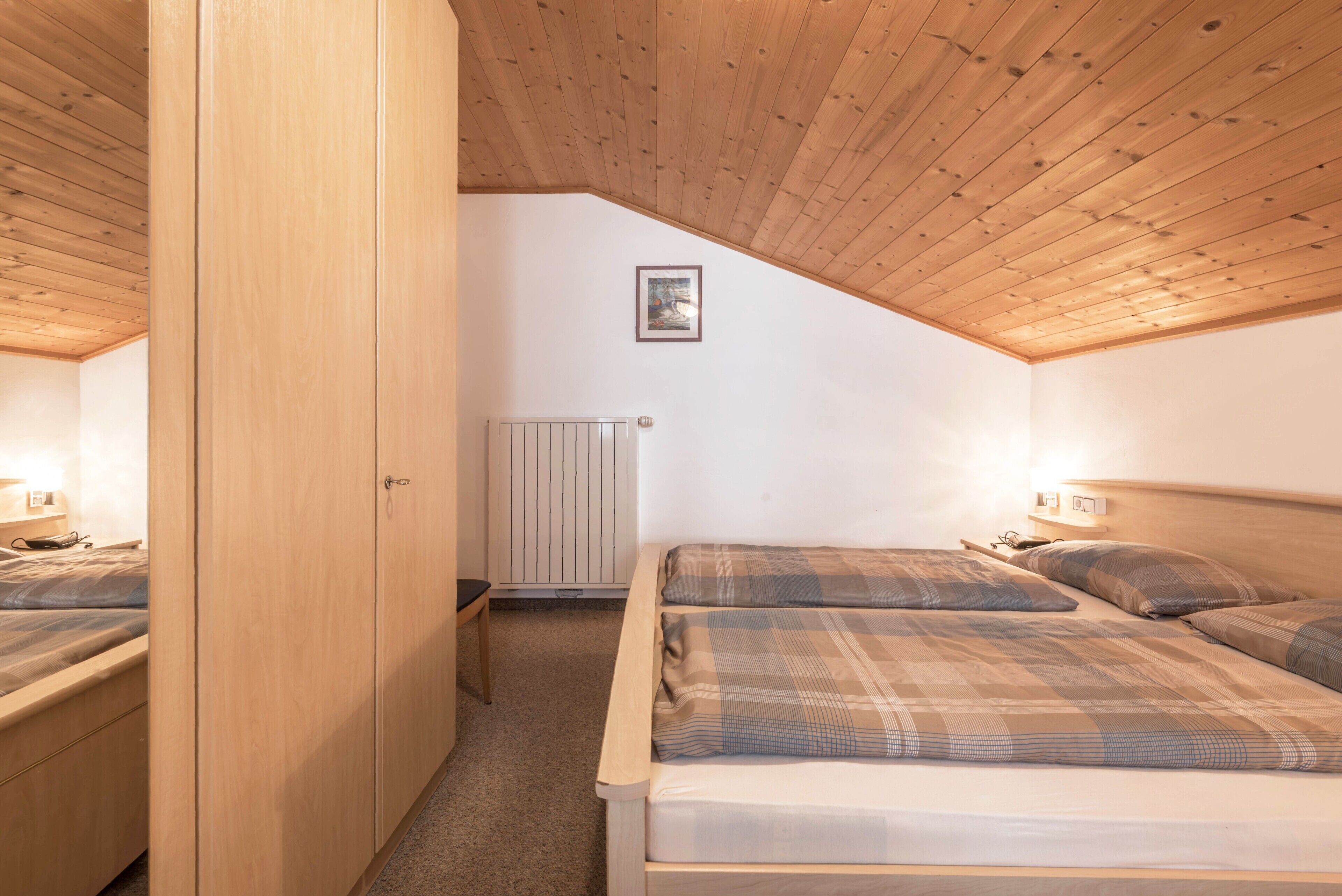 A bedroom with wooden ceiling, two beds with plaid bedding, a radiator, and a framed picture on the wall.