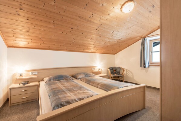 Un dormitorio con techo de madera, una cama con cabecera, mesitas de noche, una silla y una ventana con cortinas.