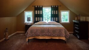 Un dormitorio con techo inclinado, una cama con ropa de cama estampada, dos ventanas con cortinas, una mesita de noche con lámpara, un aparador y una pequeña decoración de faro.