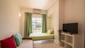 A small, well-lit room with a bed, a TV on a stand, a plant, and a window with curtains. - Beautiful Marjoh AL Holiday Apartment with Pool, Balcony, Air Conditioning and Wi-Fi (Vilamoura, Quarteira)