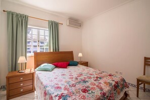 A bedroom with a bed, two bedside tables, a window with curtains, an air conditioning unit, and a chair. - Beautiful holiday apartment Marjoh AL with pool, balcony, air conditioning and Wi-Fi (Vilamoura, Quarteira)