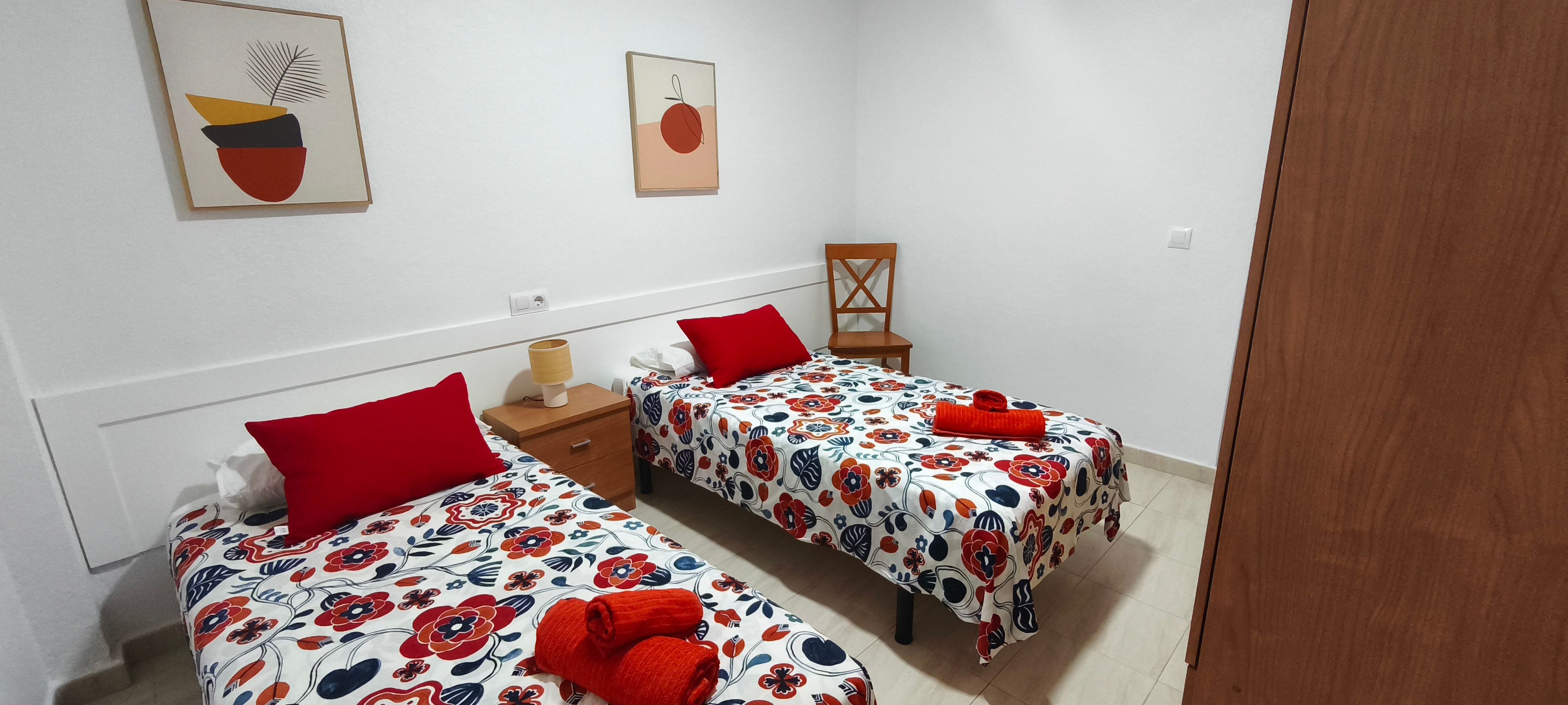 A hotel room with two beds, each with a red pillow and a floral bedspread. There is a wooden nightstand with a lamp, a chair, and a small table with a red towel.