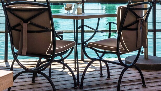 A wooden deck with two chairs and a table under a yellow umbrella, overlooking a body of water with a lighthouse and statue in the background. - High-Quality Apartment “Ferienwohnung Steinhilber 1” (Eriskirch)