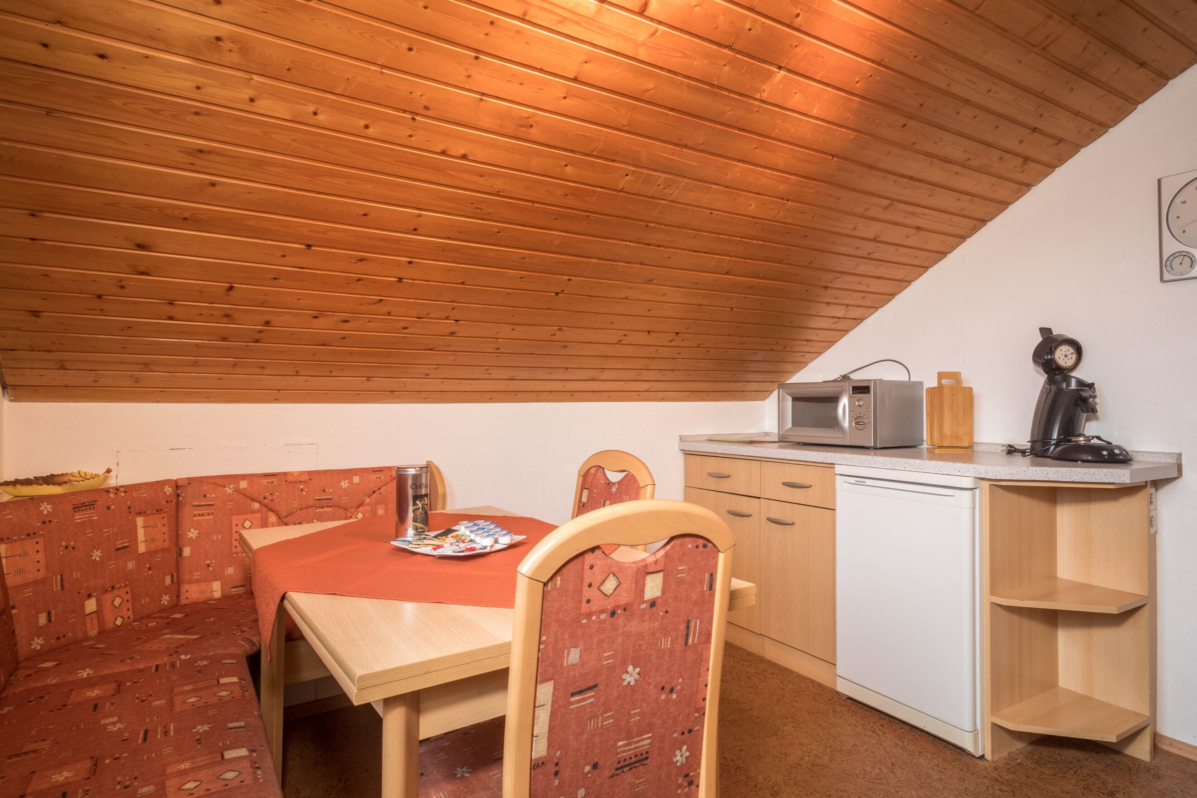 A compact kitchen with a wooden ceiling, a dining table, chairs, a microwave, a kettle, and a dishwasher.