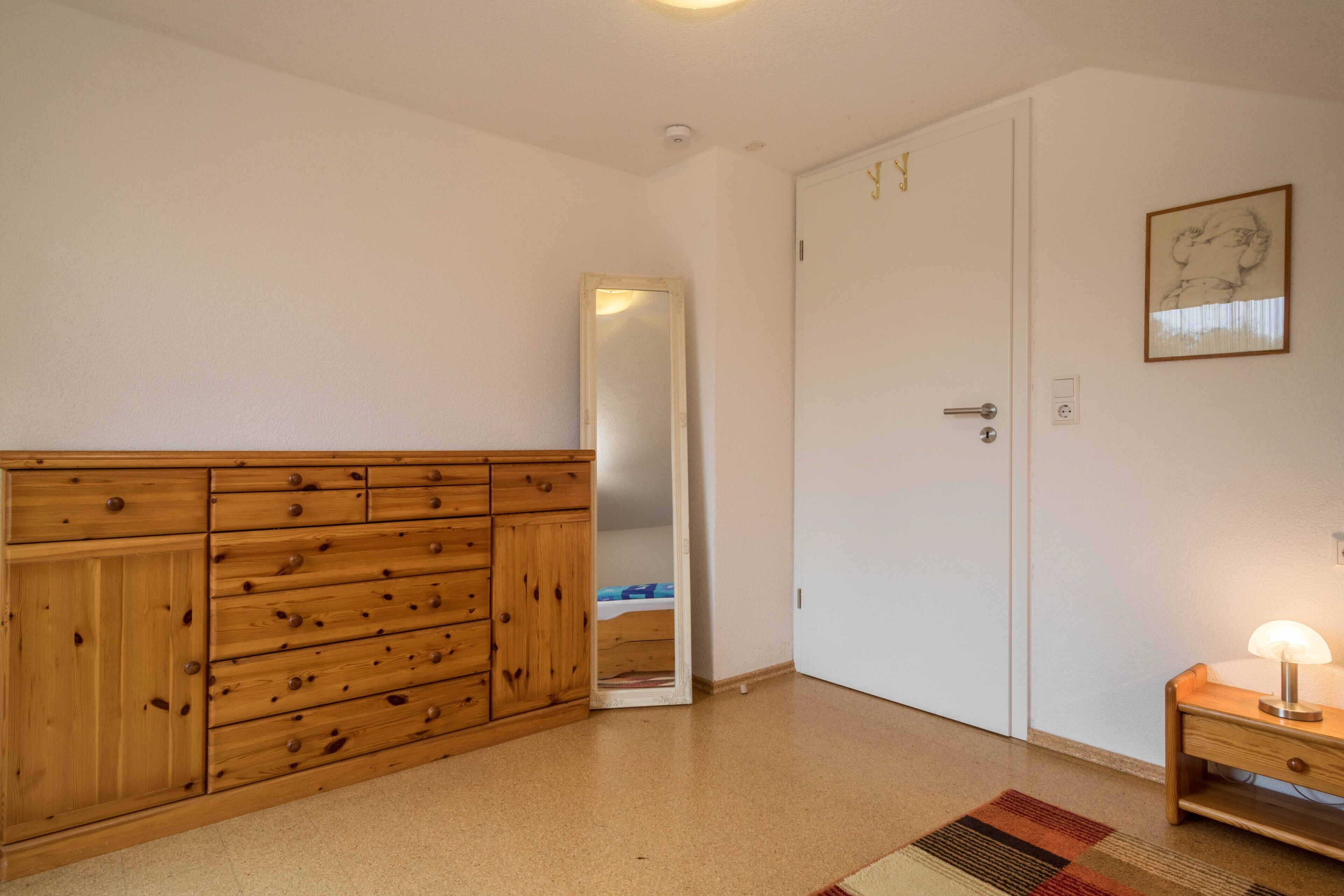 Een kamer met een houten dressoir, een deur, een spiegel, een nachtkastje met een lamp en een kamerbreed tapijt met een patroon.