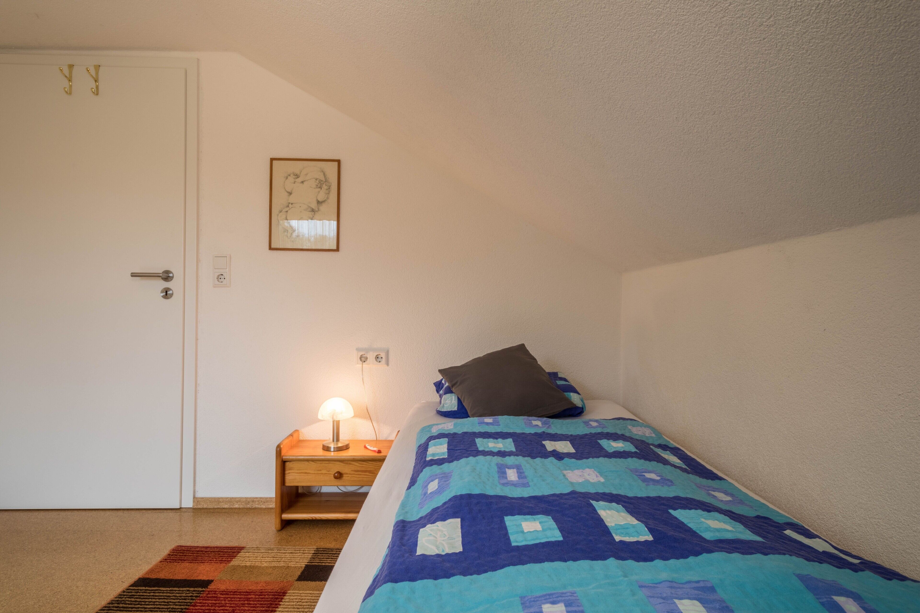 A bedroom with a bed, a nightstand with a lamp, a door, and a framed picture on the wall.