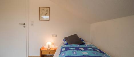 A bedroom with a bed, a nightstand with a lamp, a door, and a framed picture on the wall.