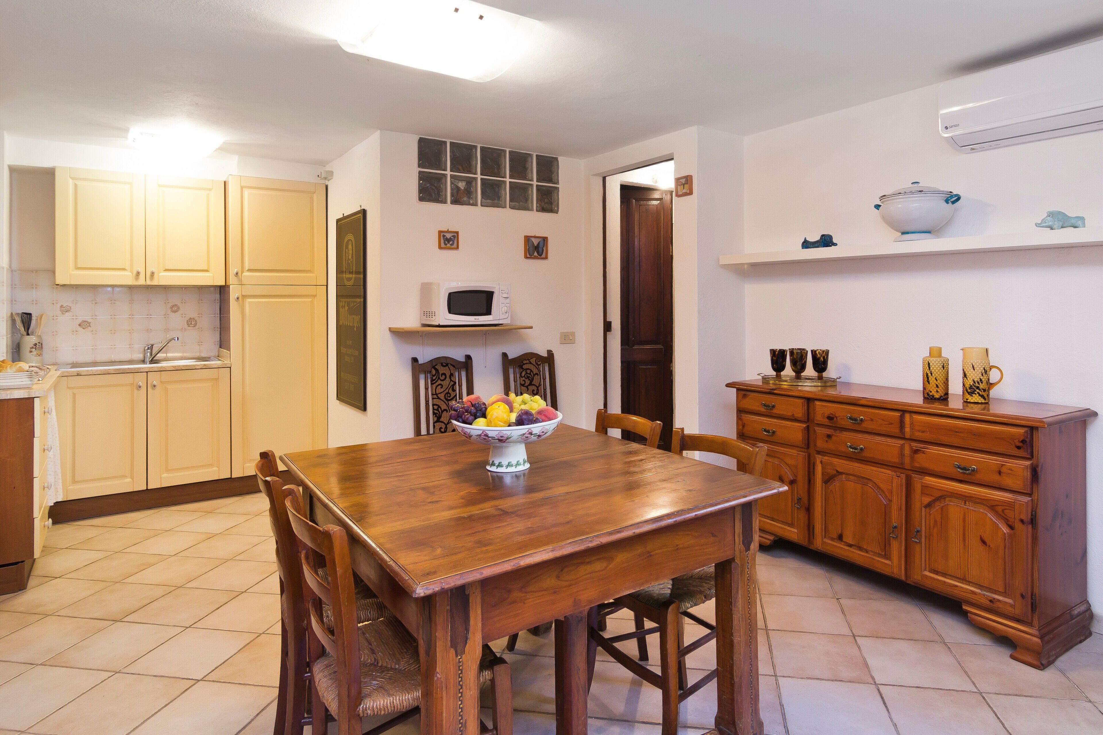 A kitchen with a wooden table, chairs, a fruit bowl, a microwave, and wooden cabinets.
