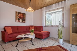 En stue med en rød sofa, et hvidt sofabord, en hængende lampe, et vindue med lette gardiner og en potteplante.