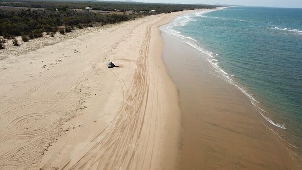 A wide sandy beach with gentle waves, a clear sky, and sparse vegetation.