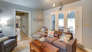 A living room with a sofa, ottoman, and coffee table, a dining area with chairs, and a kitchen in the background. - Walk to Seaside Beach! House w/ Hot Tub & Deck (Seaside)