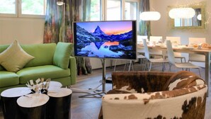 A modern living room with a green sofa, a large flat-screen TV, a dining area with a table and chairs, and a large window with curtains.