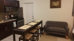A compact kitchen with dark wooden cabinets, a microwave, and a stove. There is a dining table with four chairs and a gray sofa in the living area. - Cozy Villa in Nassau with homely feel in quiet neighborhood.  (Nassau)