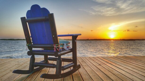 A rocking chair on a wooden deck overlooking a body of water with a sunset in the background.