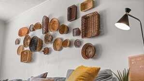 Een kamer met een grijze bank, een geel kussen en een muur versierd met diverse gevlochten manden en een lamp.
