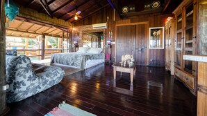 A spacious room with a bed, a sofa, and a small table. There is a ceiling fan, wooden cabinets, and a large window.