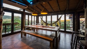 A wooden dining area with a large table and benches, large windows with a view of greenery, and hanging pendant lights.