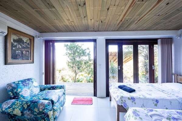 Une pièce avec un plafond en bois, un canapé à motifs floraux, un lit recouvert d’une courtepointe à fleurs et une vue sur la verdure à l’extérieur.