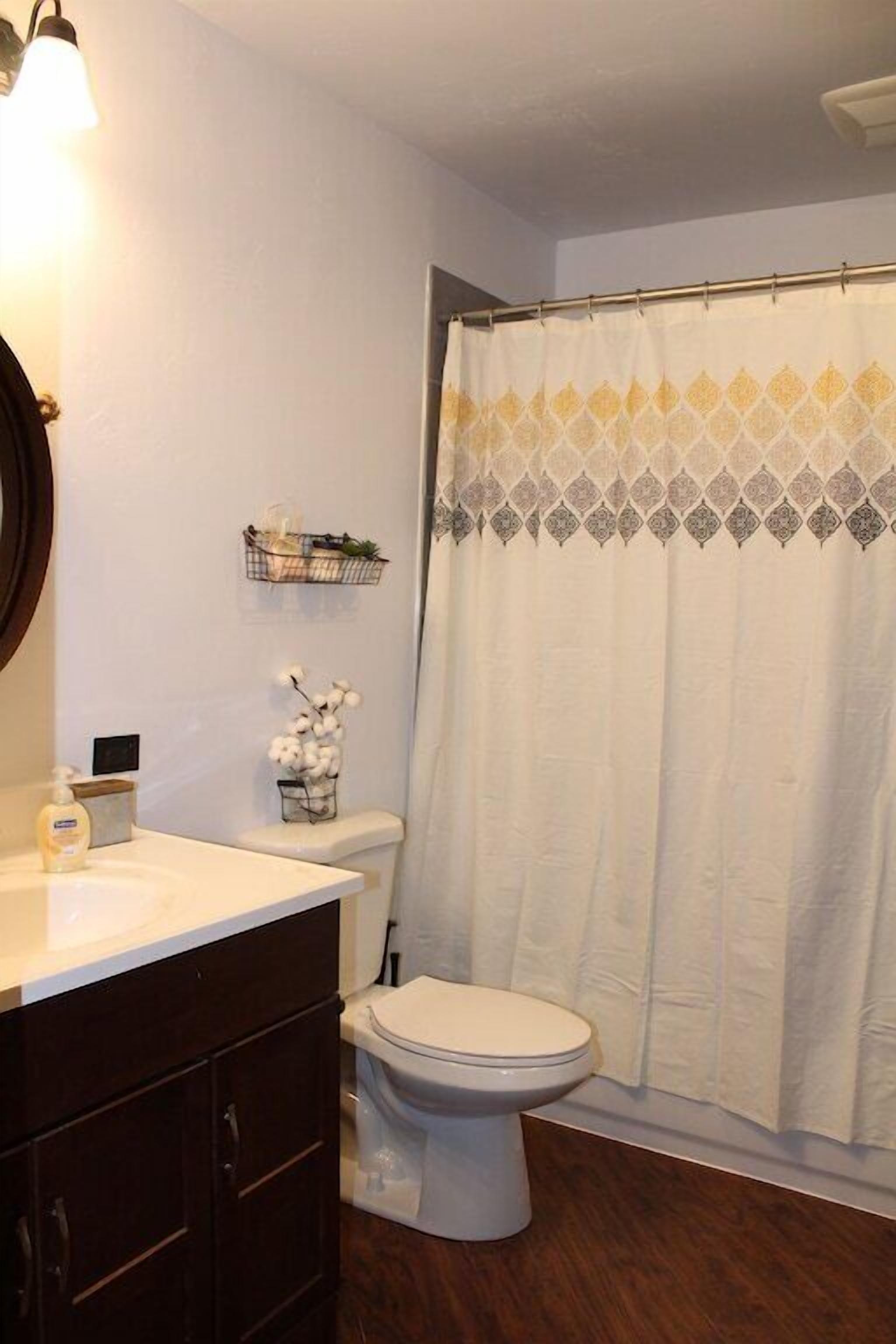 A bathroom with a white toilet, a wooden vanity, a shower curtain with a geometric pattern, and a small shelf with toiletries.
