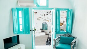 Une pièce avec un téléviseur posé sur un meuble blanc, un fauteuil turquoise et une vue sur un bâtiment blanc, visible à travers une porte dotée de volets turquoise.