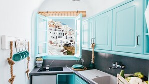 A kitchen with teal cabinets, a window with a view of white buildings, a small refrigerator, and a wooden crate with vegetables. - Blessed Blue Boutique Cave Suite in the heart of Oia (Oia)