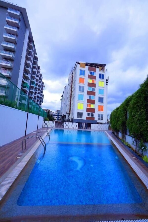 A swimming pool with a wooden deck, surrounded by modern buildings with balconies and a clear sky. - The Altruist Whitefield Hotel (Bengaluru)