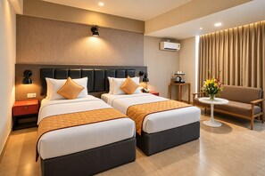 A hotel room with two beds, a sofa, a small table with a vase of flowers, an air conditioning unit, and a wall-mounted lamp.