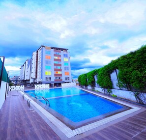 A swimming pool with a wooden deck, surrounded by a multi-story building with colorful windows and a green hedge. - The Altruist Whitefield Hotel (Bengaluru)