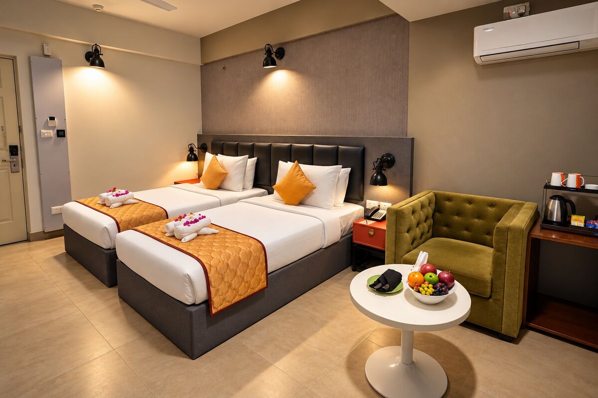 A hotel room with two beds, a small table with fruit, a green armchair, and a coffee maker.