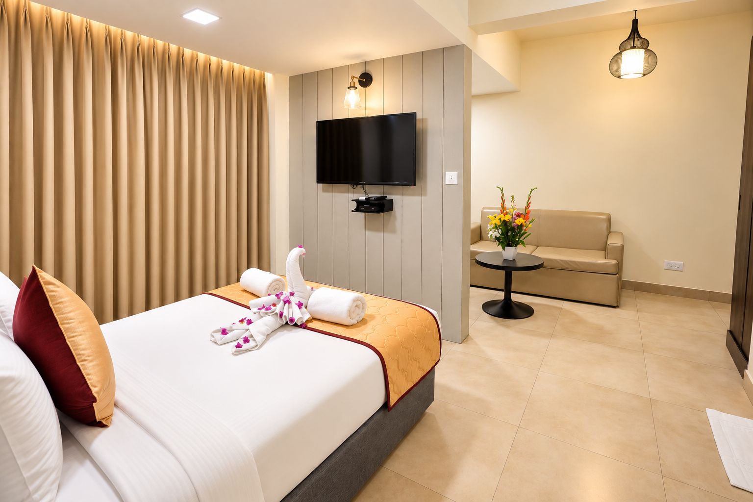A hotel room with a bed, a TV mounted on the wall, a sofa, a small round table with flowers, and a curtain.