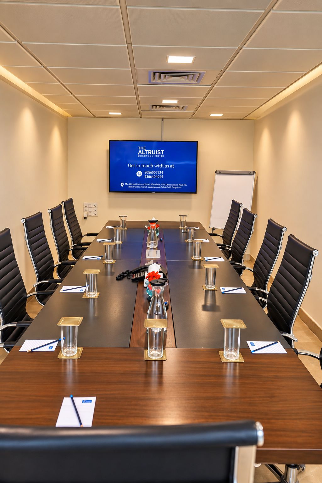 A conference room with a long table, black chairs, a whiteboard, and a screen displaying hotel information.
