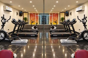 A mirrored gym with multiple treadmills and stationary bikes, a central wall with a digital display, and air conditioning units on the walls.