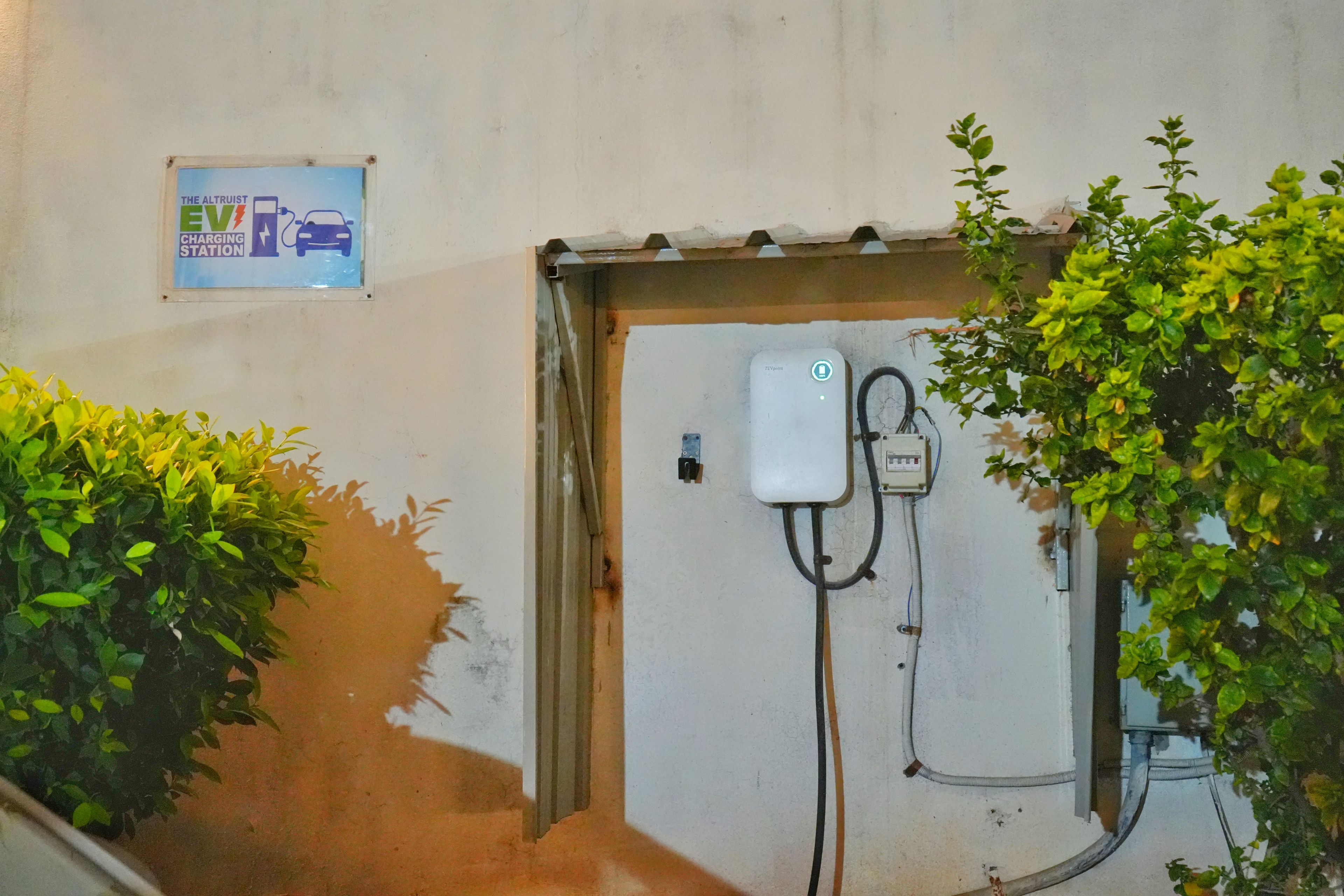 Uma estação de carregamento de veículos elétricos com sinalização acima, caixa elétrica na parede e vegetação nas laterais.