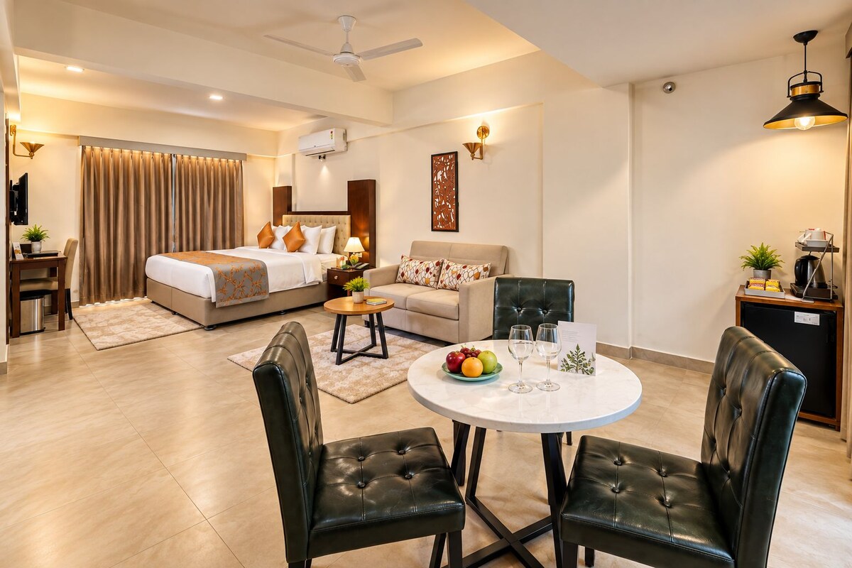 A modern hotel room with a bed, a sofa, a dining table with chairs, a ceiling fan, and a small kitchenette.