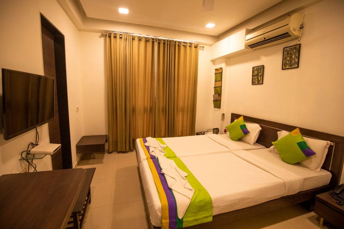 A hotel room with a large bed, a desk, a chair, an air conditioning unit, and a television.
